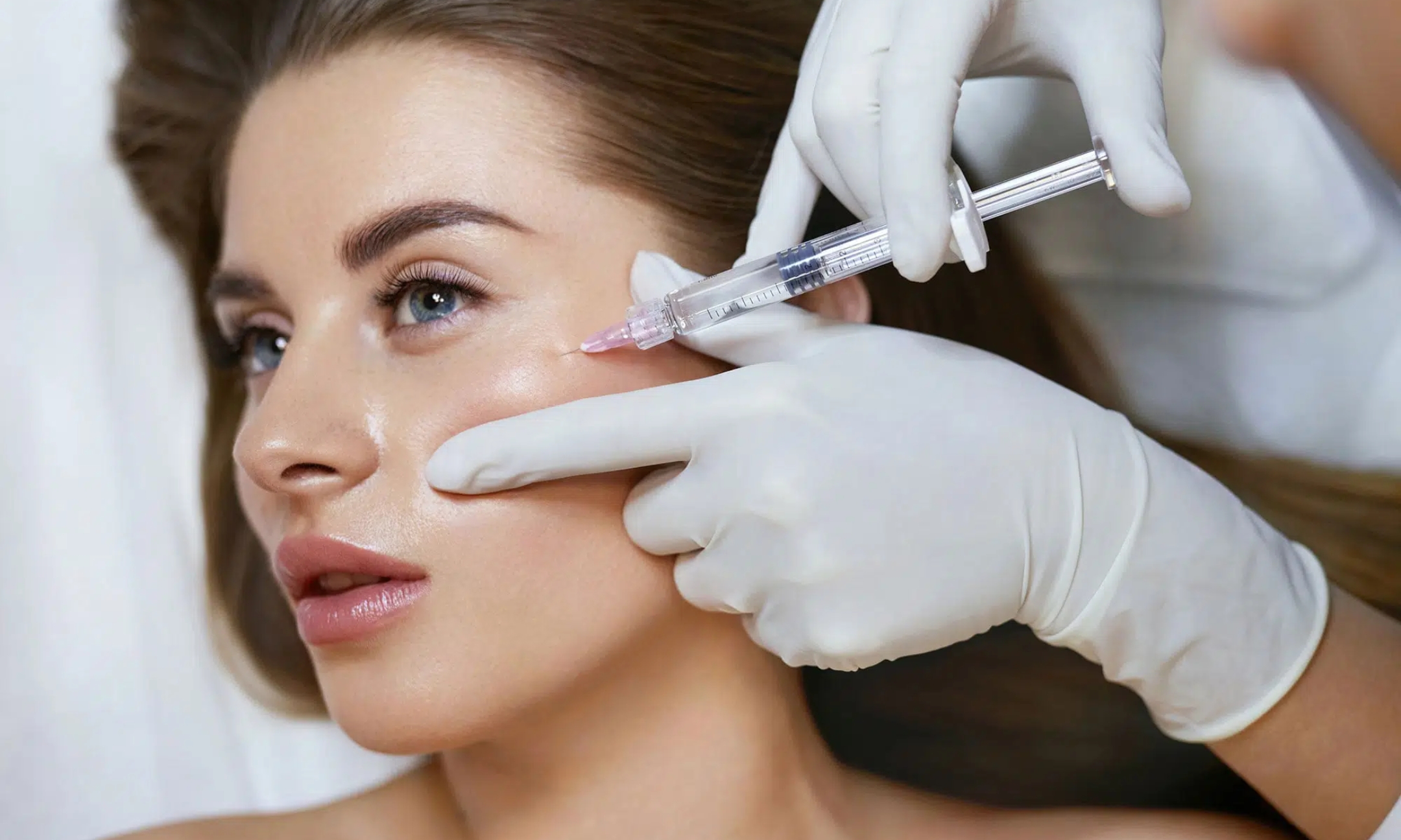 Close-up of a patient receiving a facial cosmetic injection near the cheekbone from a practitioner wearing white medical gloves.