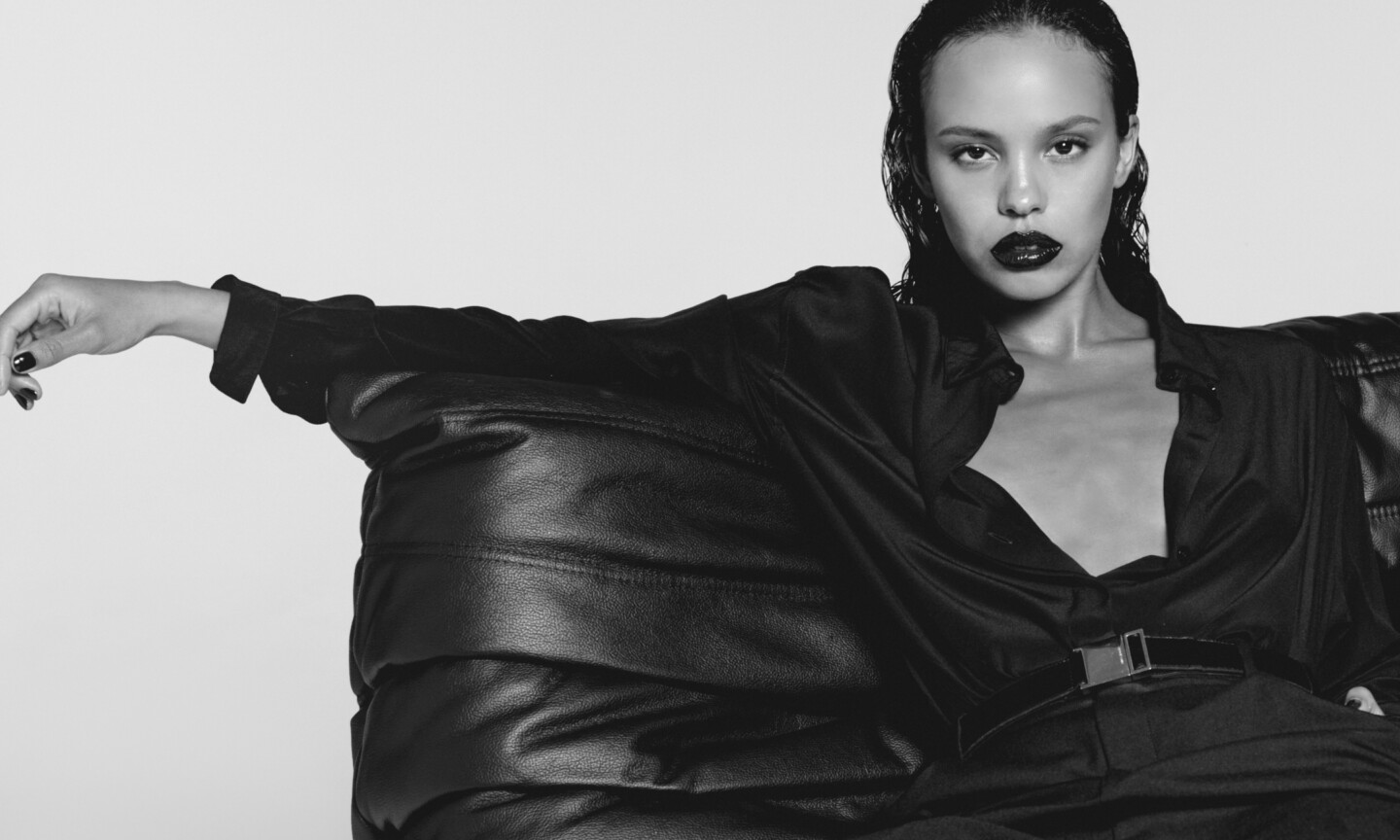 Black-and-white campaign image of actress Alisha Boe for YSL Beauty Black Opium Pink Glaze, wearing an open dark button-down shirt while leaning on a black leather sofa with dark lips and slicked-back hair.