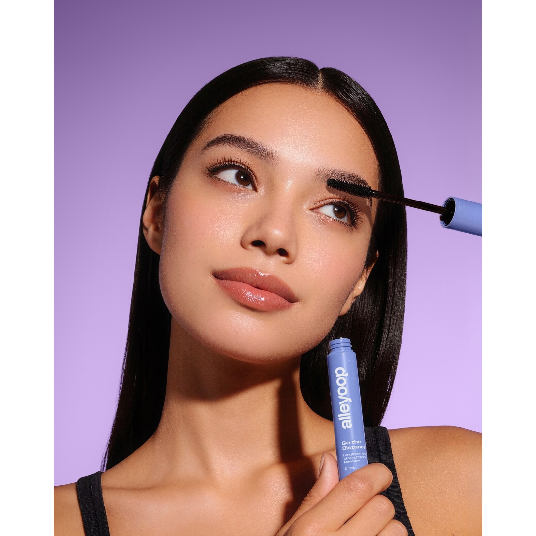 A model with long, sleek dark hair applies Alleyoop Go the Distance Lengthening & Strengthening Mascara in Black while holding the tube, against a purple backdrop