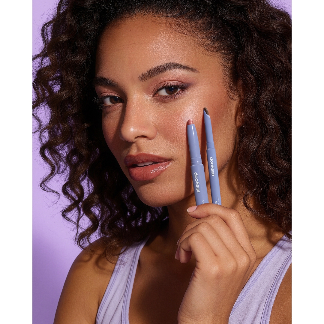 A model with curly hair holds the Alleyoop 11th Hour Cream Eyeshadow & Liner Stick and the Swift Brow Shape + Fill Brow Pencil up to her cheek, against a lilac backdrop