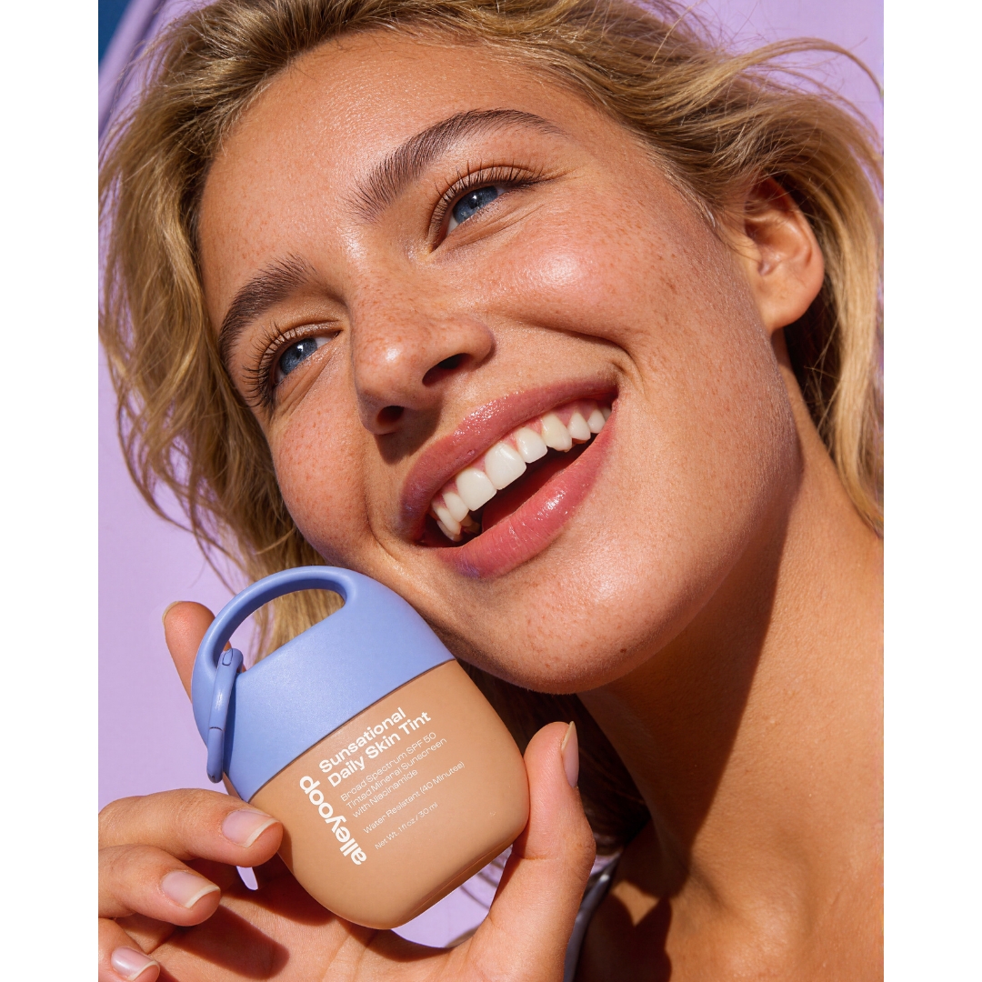 A smiling model with blonde hair and freckles holds Alleyoop Sunsational Daily Skin Tint SPF 50 to her cheek in bright sunlight