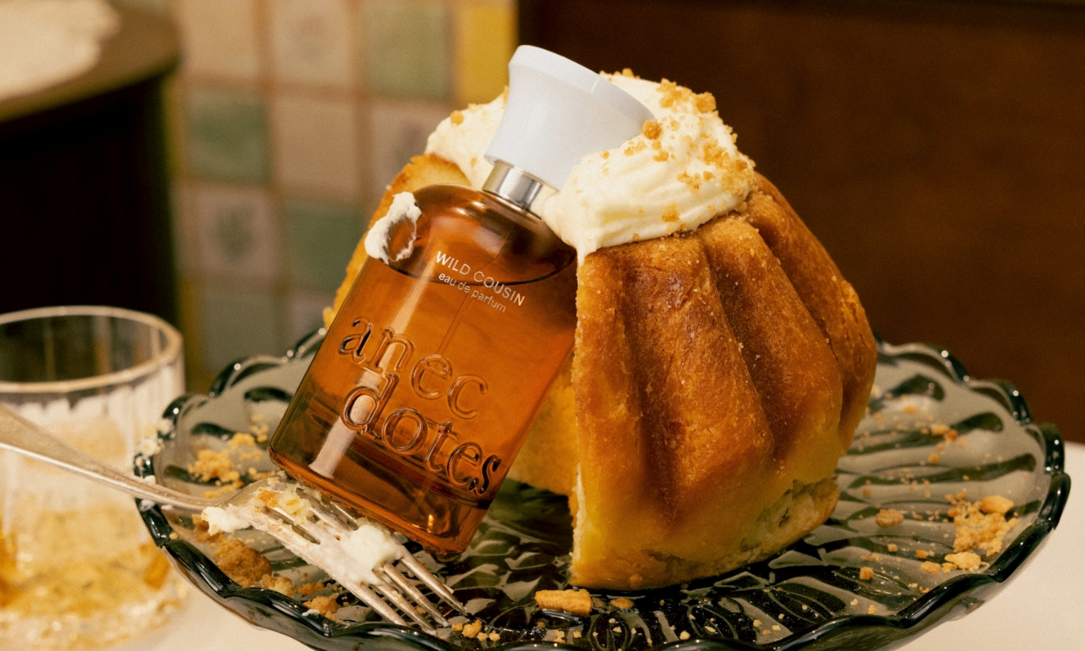 An amber glass bottle of Anecdotes by Artists Wild Cousin Eau de Parfum leaning against a slice of cake topped with whipped cream on a glass cake stand.