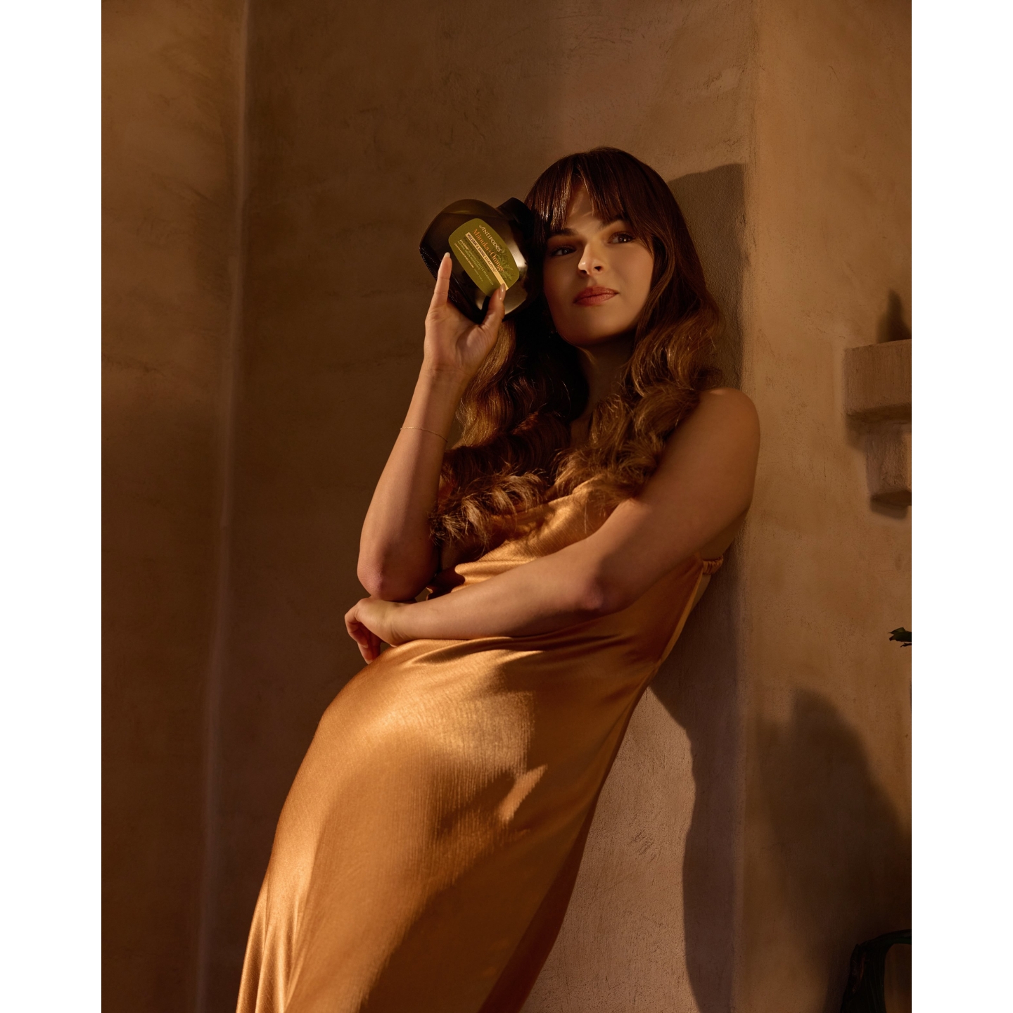 A model in a silky gold slip dress leaning against a textured wall while holding an Antipodes hair product, featured in the brand's natural haircare launch announcement on Beauty News Daily.