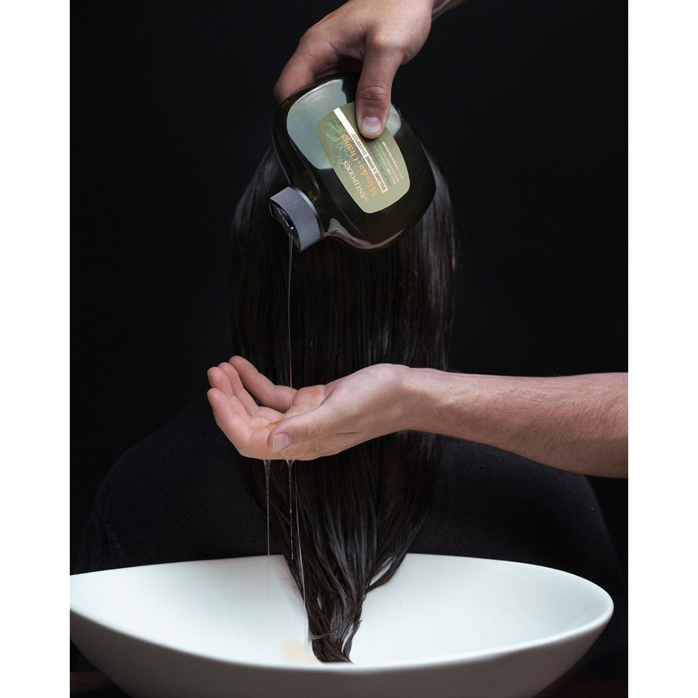 Close-up of a dark-green bottle of Antipodes Mānuka and Orange Volume and Shine Shampoo being poured over a model's wet hair over a white basin, featured on Beauty News Daily.