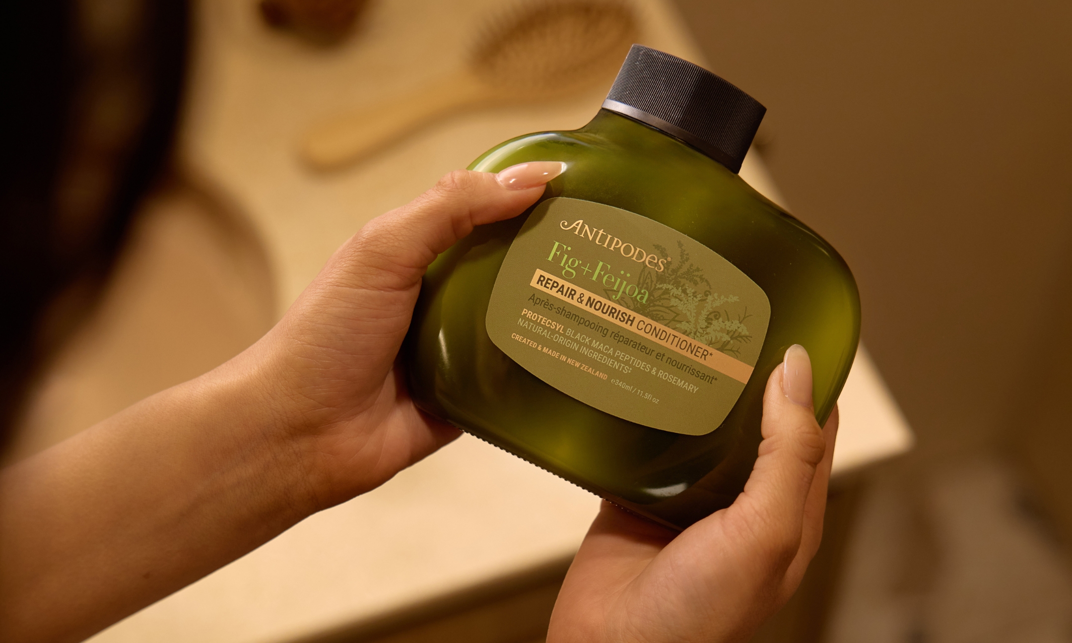 A close-up of hands holding a dark-green bottle of Antipodes Fig and Feijoa Repair and Nourish Conditioner in a bathroom setting, featured on Beauty News Daily.