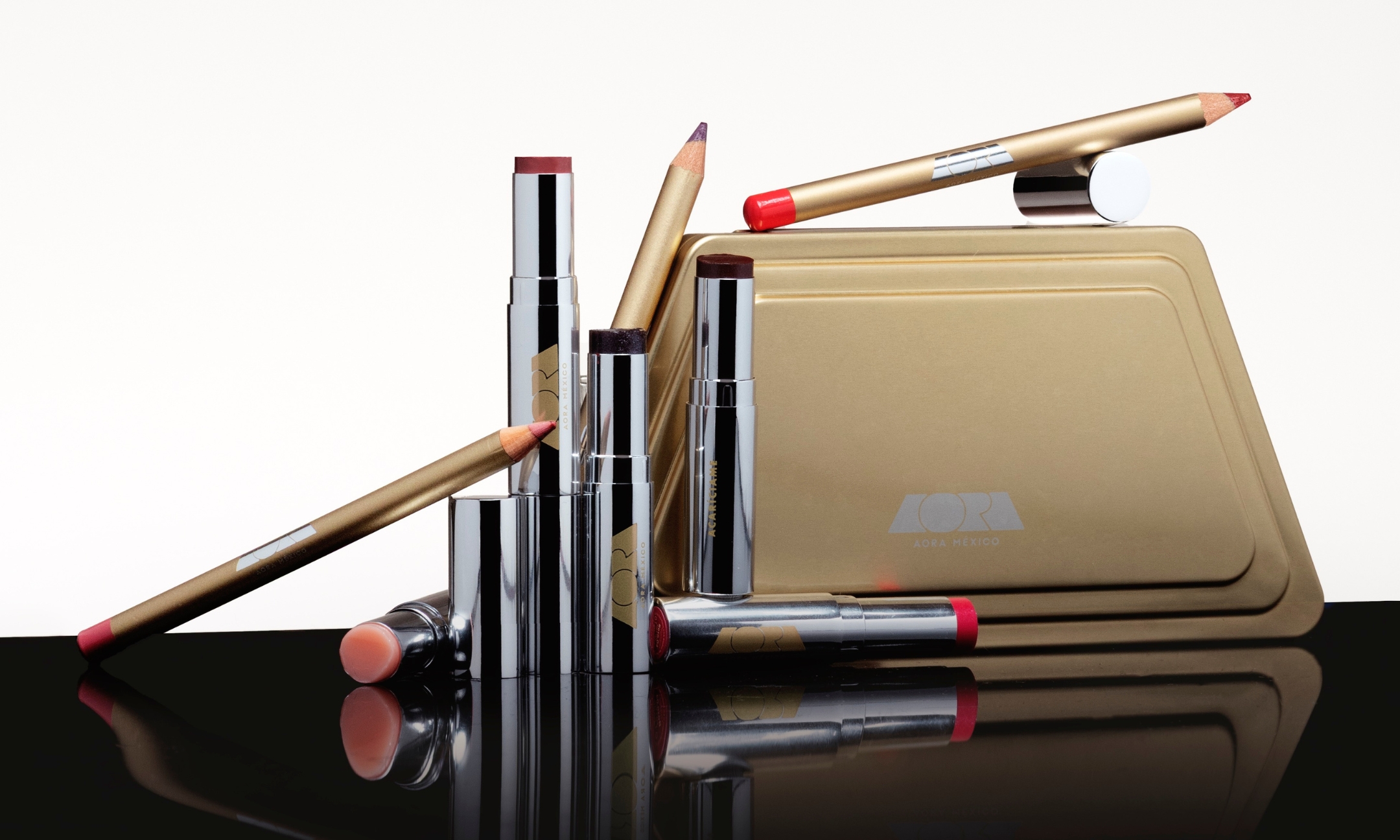 An assortment of Aora México lipsticks and eye pencils arranged next to a gold metal palette case on a reflective surface, featured in the brand's Sephora launch announcement on Beauty News Daily.