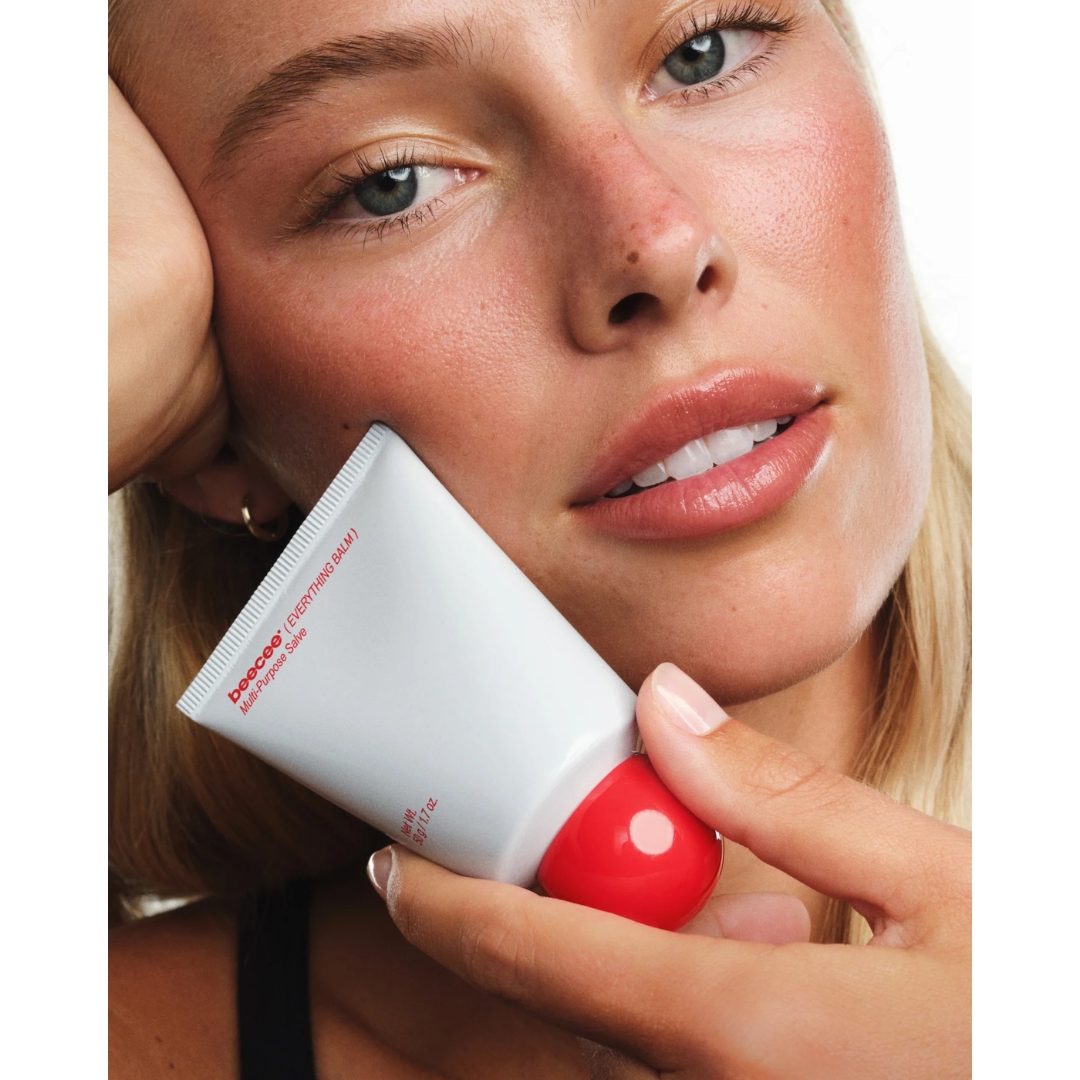 Close-up portrait of a blonde model with rosy cheeks holding a white tube of Beecee Everything Balm Multi-Purpose Salve with a red rounded cap near her jawline.