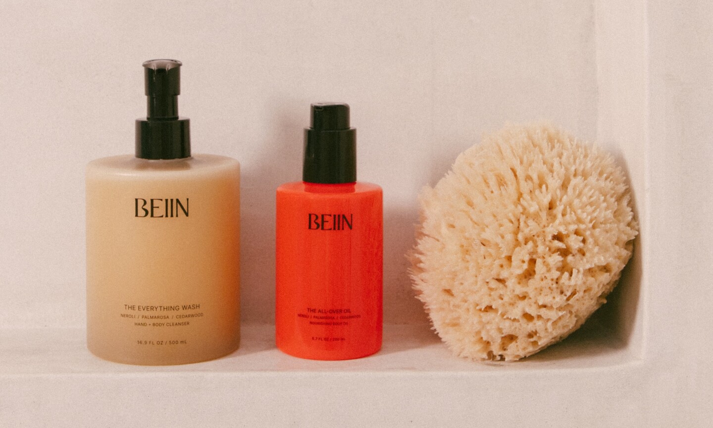 Beiin The Everything Wash and The All-Over Oil on a shower shelf next to a natural sea sponge