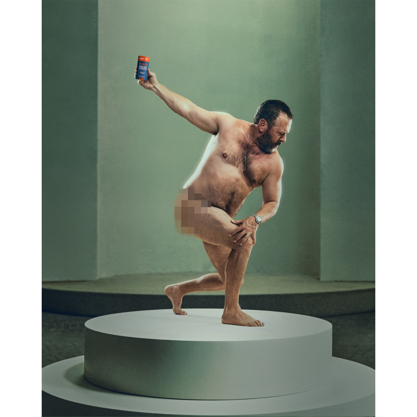 Comedian Bert Kreischer posing nearly nude on a white pedestal while holding up a blue and orange tube of Mando Pro Sport Solid Stick Whole Body Deodorant, in a tongue-in-cheek campaign shot, featured in a daily beauty news roundup on Beauty News Daily.
