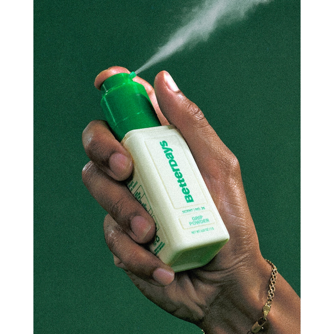 A hand pressing the green nozzle of a white BetterDays Grip Powder bottle, releasing a fine spray against a dark green background.