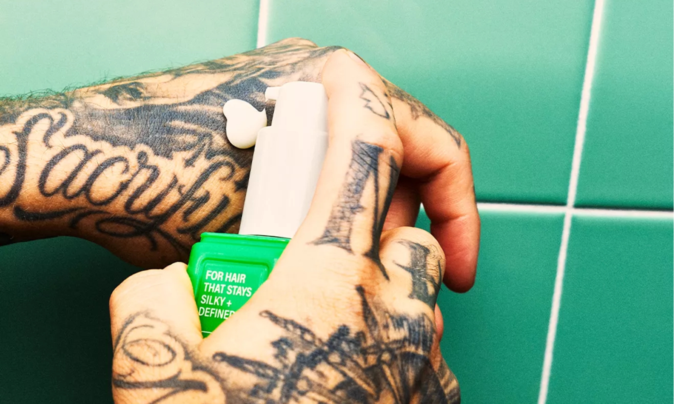 Tattooed hands dispensing a pump of BetterDays Rebound Cream onto the back of a hand against a green tiled background.