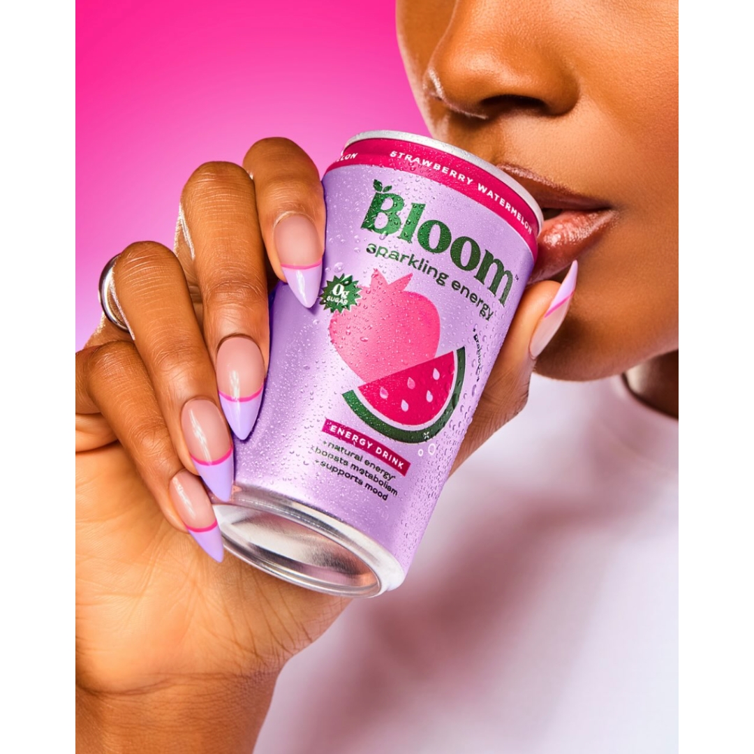 Close-up of a woman with lilac French manicure nails holding a chilled can of Bloom Sparkling Energy Strawberry Watermelon drink up to her lips against a pink background.