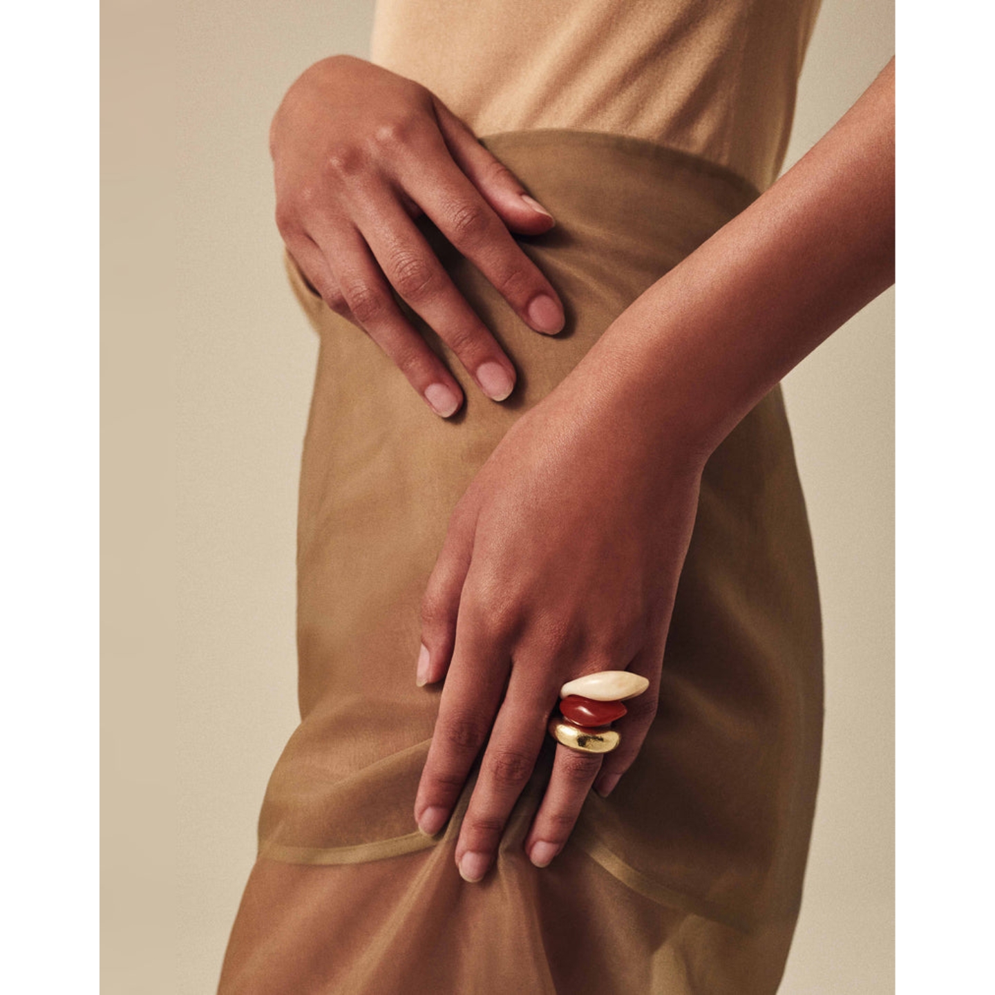 A Buff Beauty model resting clean, manicured hands over a beige outfit, showcasing natural bare nails and three stacked chunky rings in cream, terracotta, and gold.