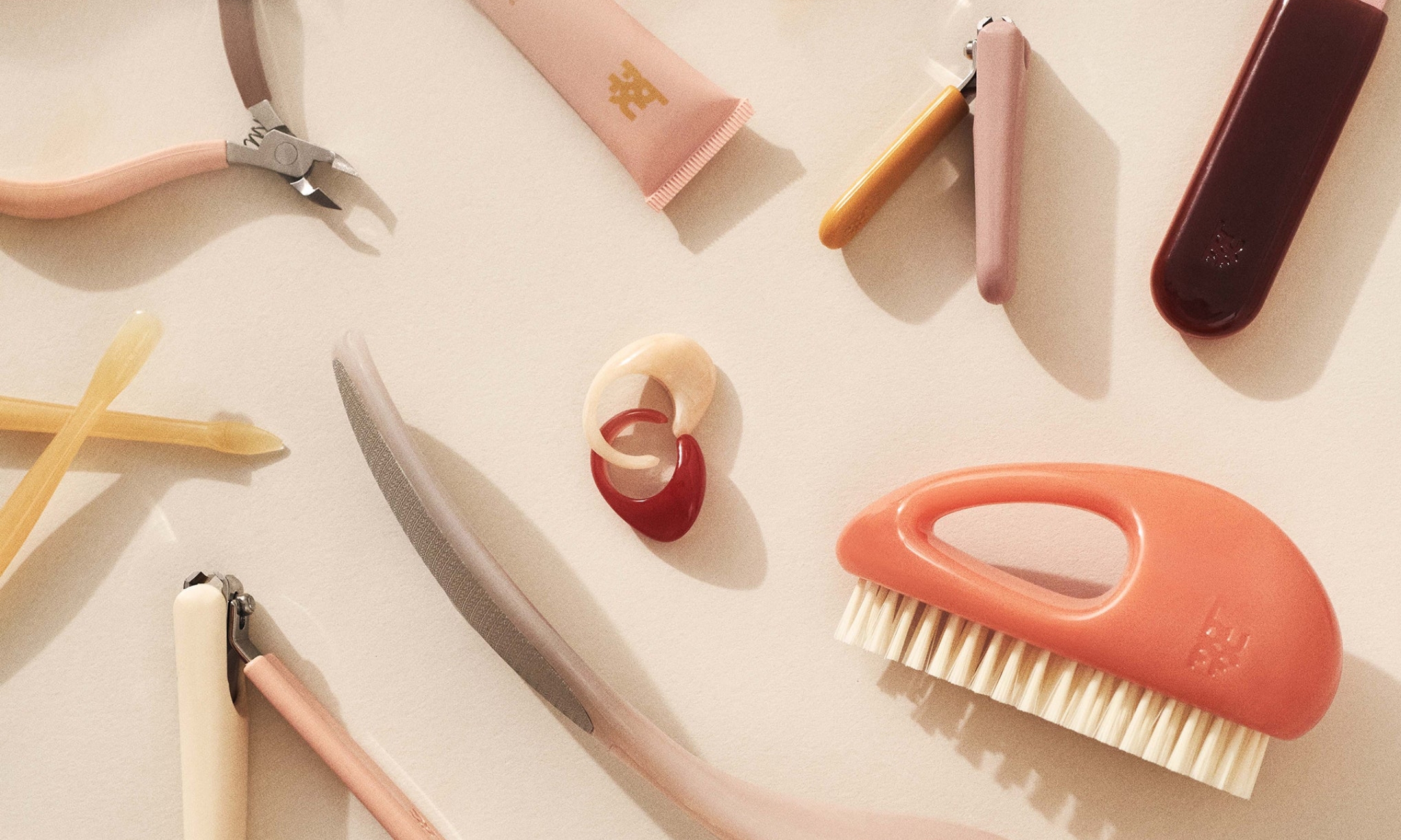 A flat lay array of modern Buff Beauty manicure tools and hand care essentials, including a hand brush, cuticle nippers, nail clippers, nail file, and gel oil tube on a warm neutral backdrop.