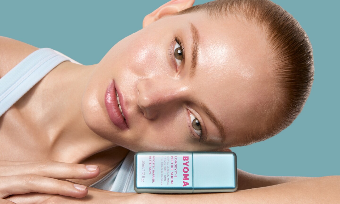Model with slicked-back hair resting her cheek on a metallic blue Byoma Longevit-8 Peptide Serum bottle against a light teal background.