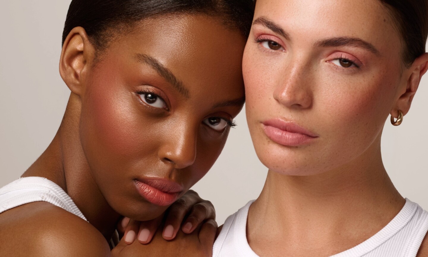 Two models posing side-by-side in white tops, showcasing a full makeup look using Cassie beauty products applied on cheeks, eyes, and lips across different skin tones.