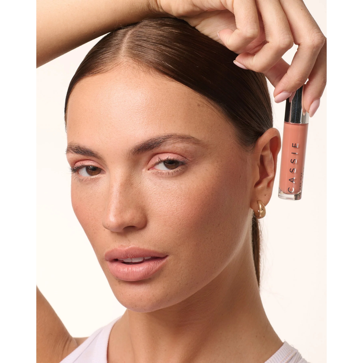 A model with sleek hair pulled back and pink eye makeup holding a Cassie We Are One Colour Wand next to her face against a plain light background.