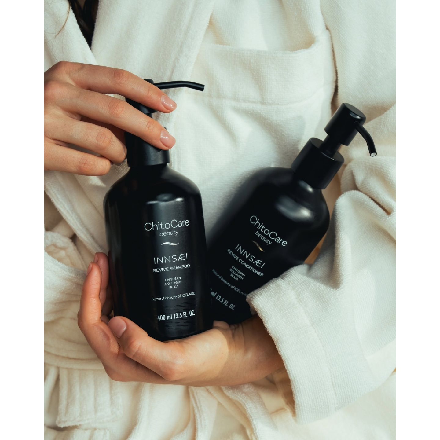 A close-up of a person in a white bathrobe holding black pump bottles of ChitoCare Beauty Innsæi Revive Shampoo and Revive Conditioner, featured on Beauty News Daily