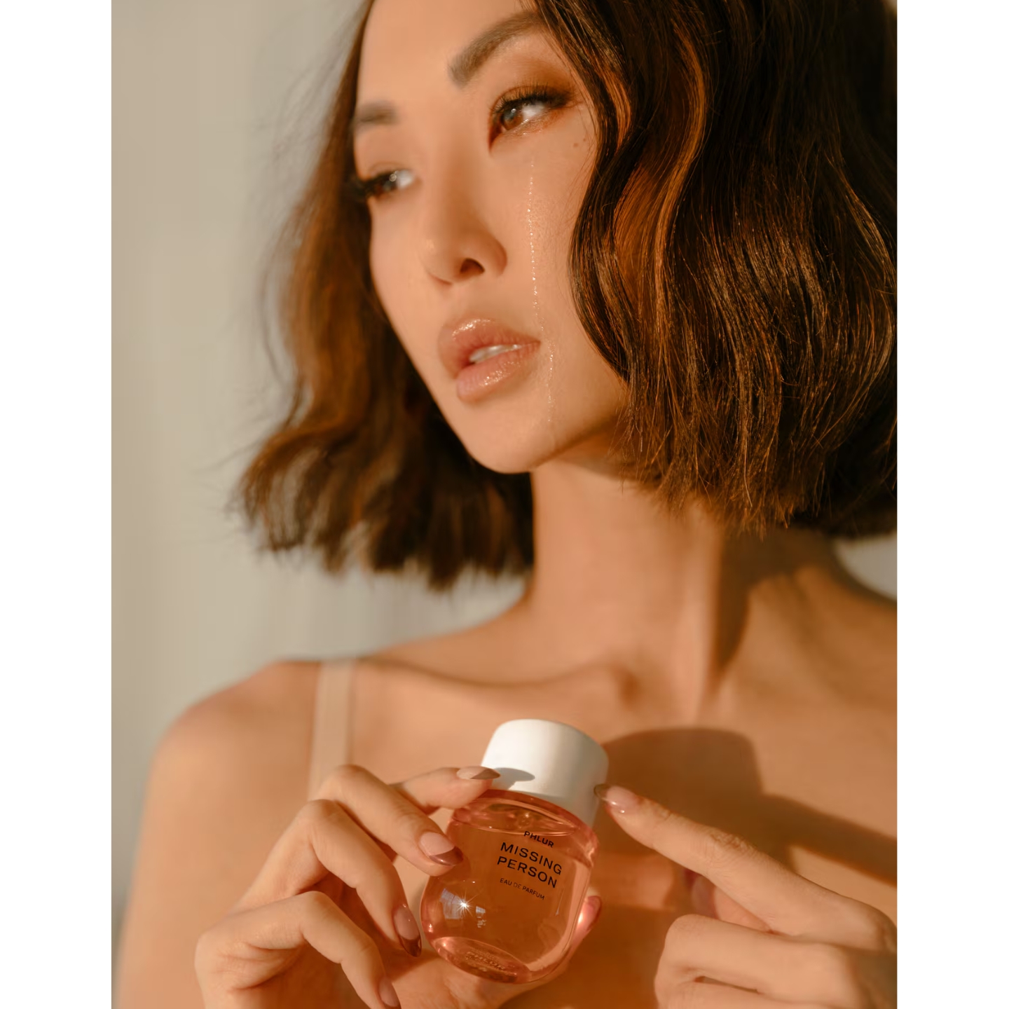 Phlur owner Chriselle Lim with a single tear on her cheek holding a bottle of Phlur Missing Person Eau de Parfum.