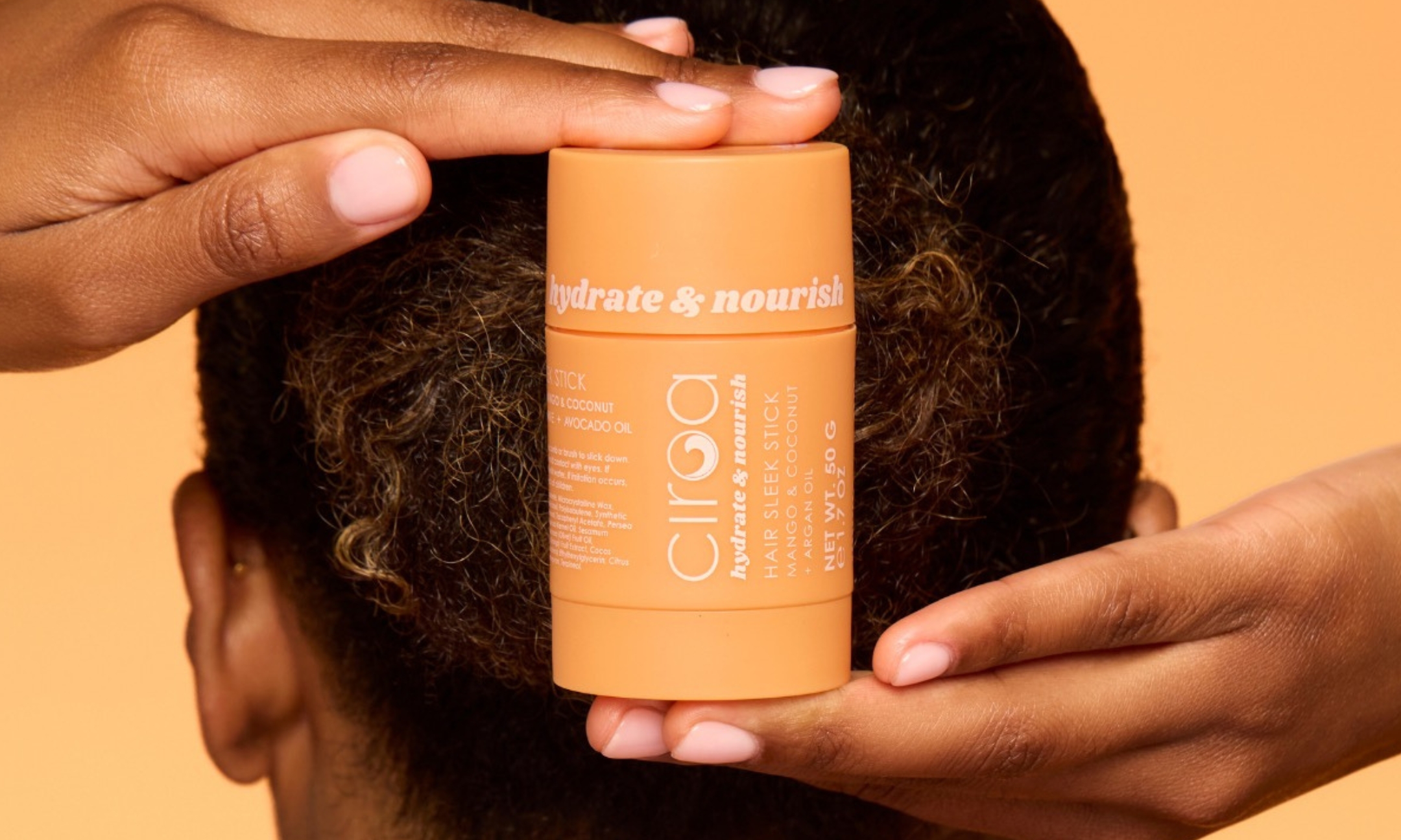 Close up of hands holding the orange Ciroa Hydrate and Nourish Hair Sleek Stick bottle against a sleek hairstyle, featured on Beauty News Daily.