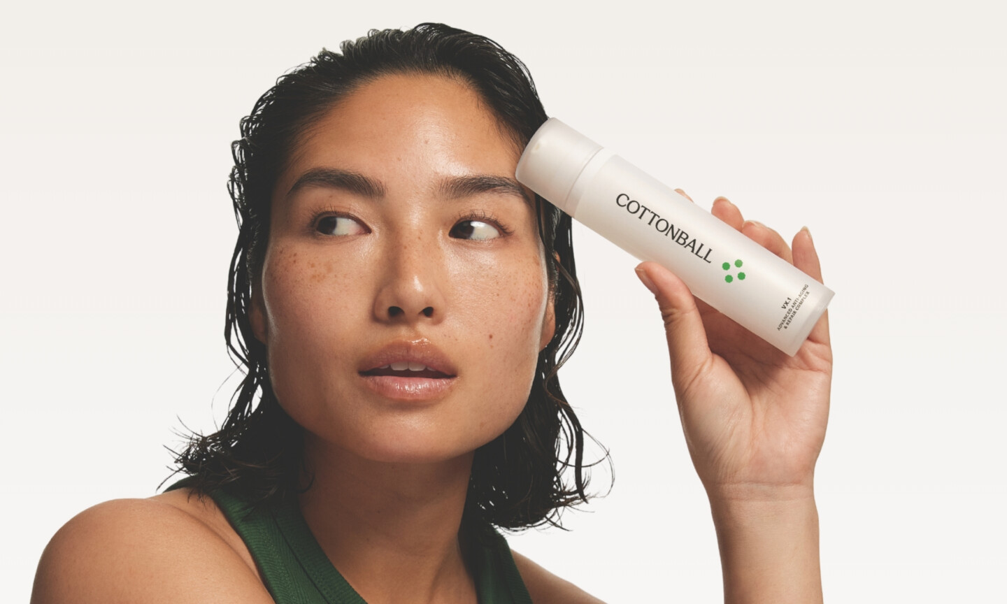Model with damp hair holding a frosted white bottle of Cottonball VX.1 Advanced Anti-Aging & Repair Complex up to her forehead against a white studio background.