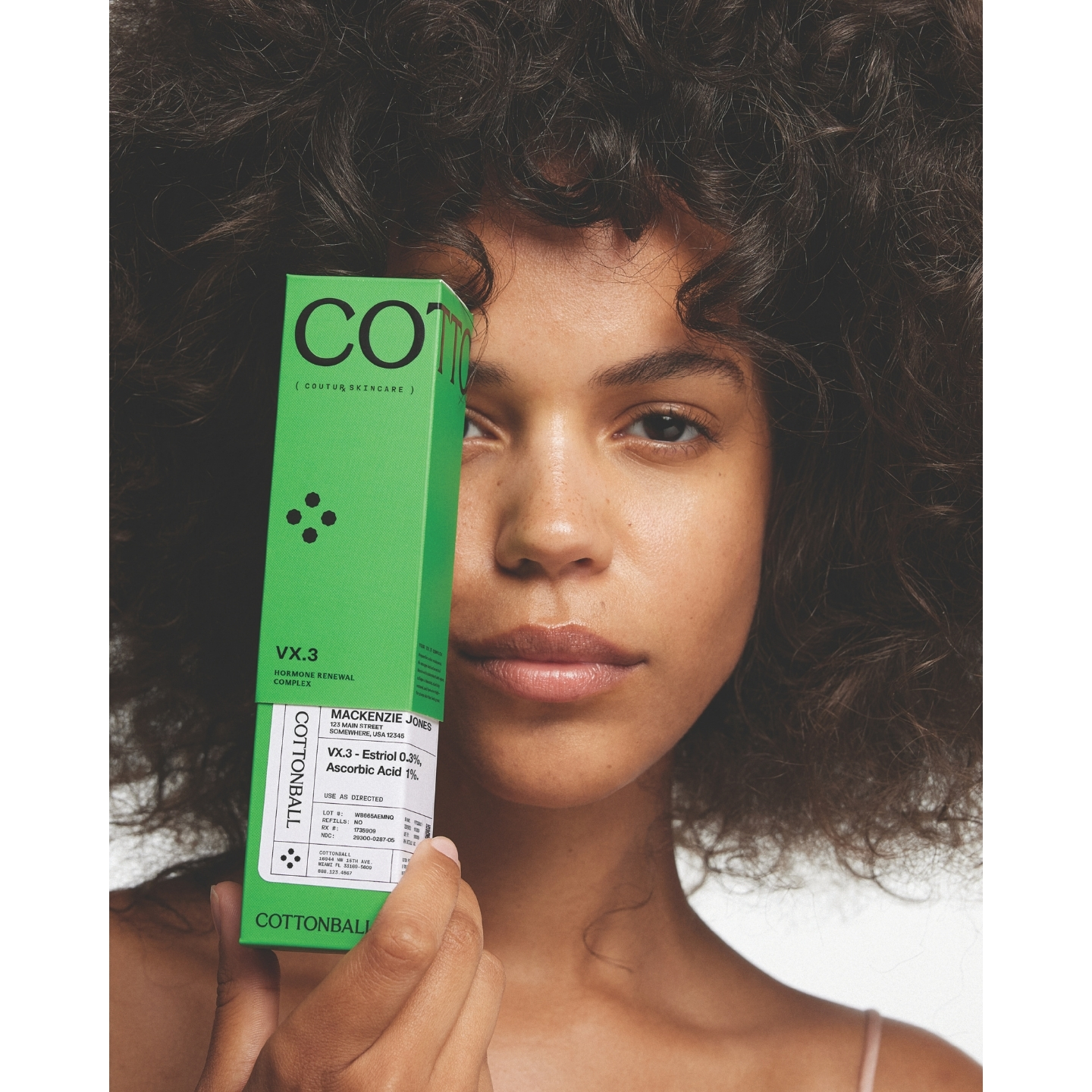 Model with curly dark hair holding a bright green box of Cottonball VX.3 Hormone Renewal Complex in front of half her face against a plain white background.
