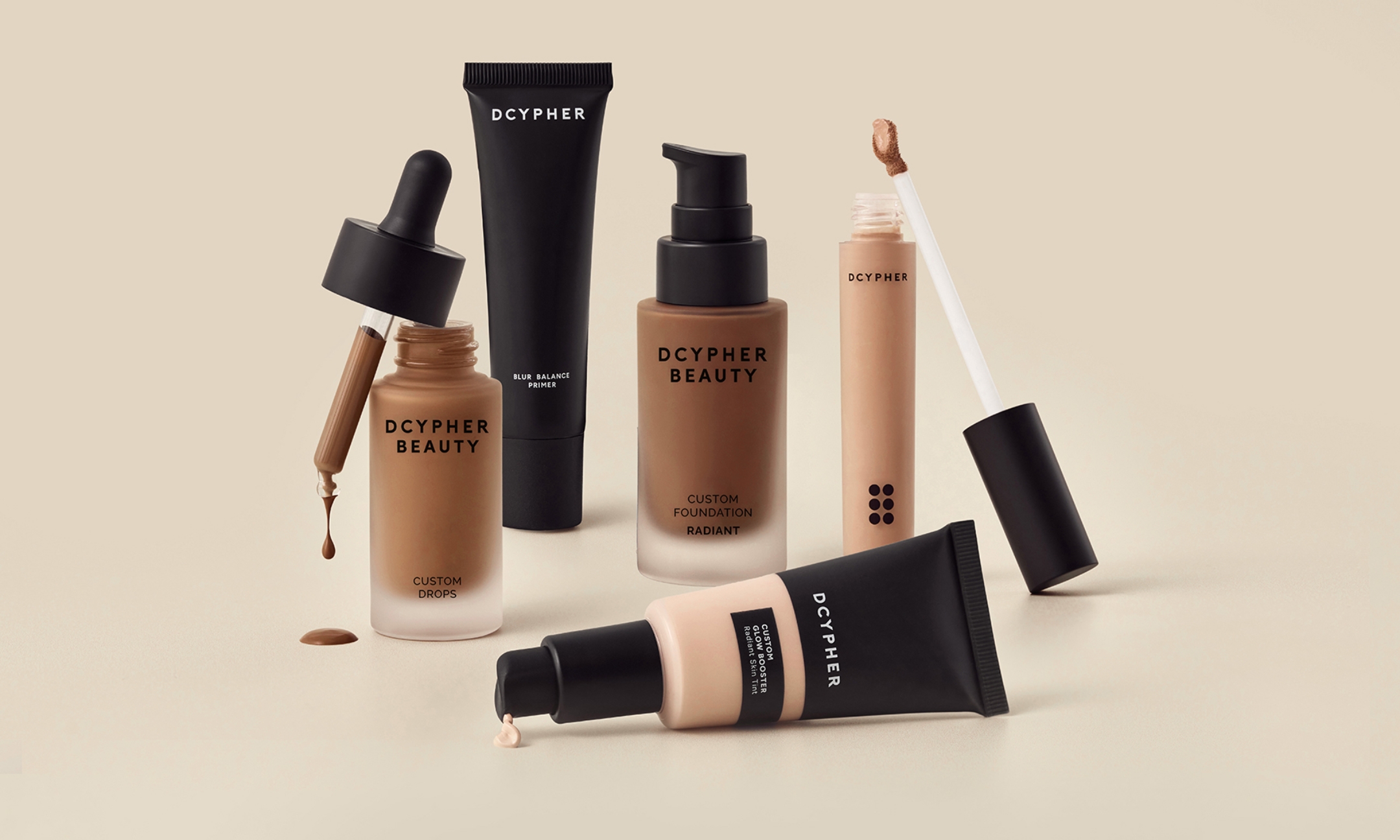 An assortment of DCYPHER custom complexion makeup products including Custom Drops, Custom Foundation, and Custom Glow Booster in black and frosted glass packaging, featured in the brand's John Lewis UK launch announcement on Beauty News Daily.