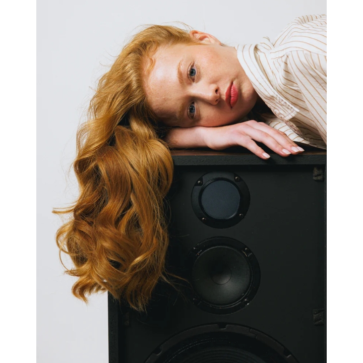 A model with long, wavy red hair resting her head on a large black audio speaker, featured in the Decibel haircare UK launch announcement on Beauty News Daily.