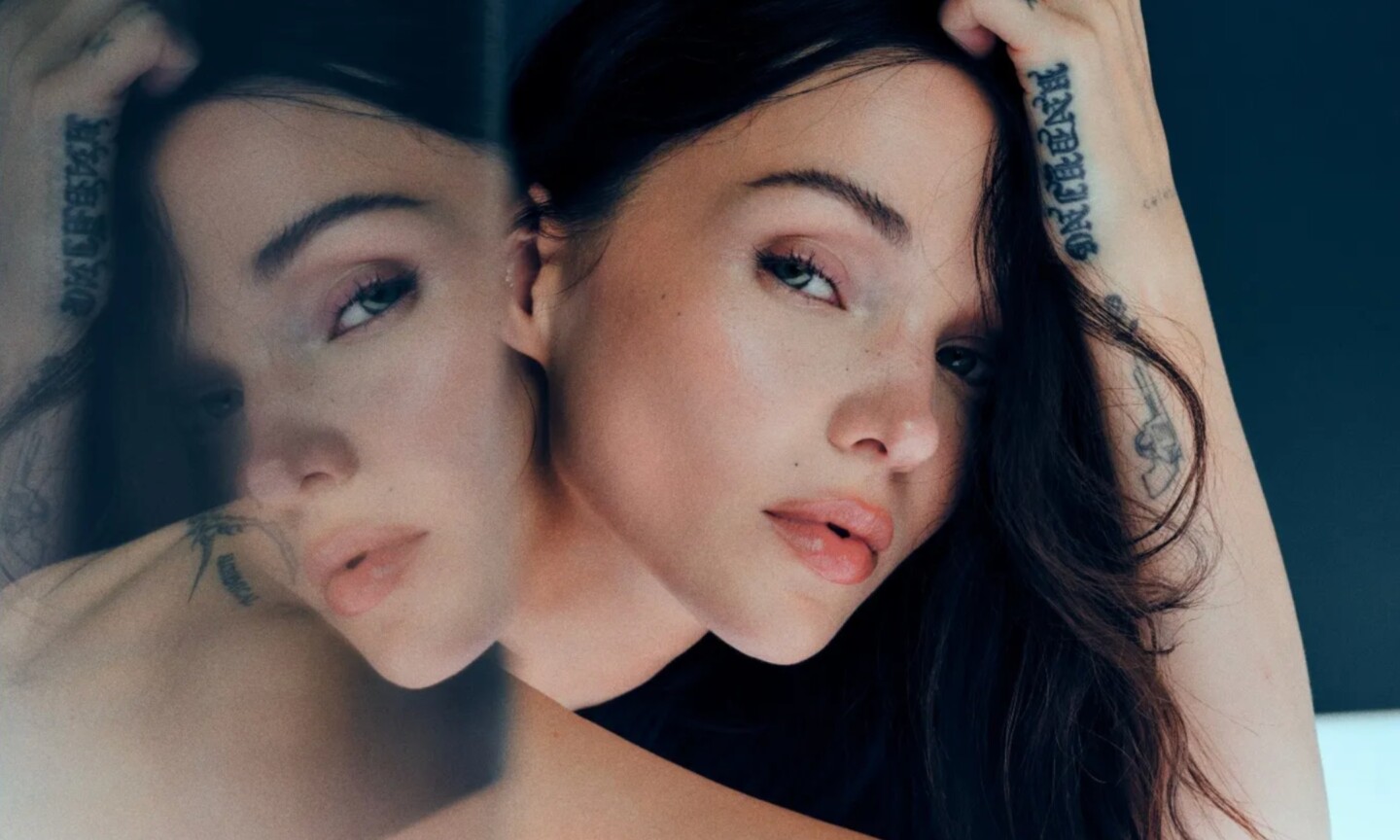 Dove Cameron, with glowing skin and tattoos on her arm, poses with one hand in her dark hair, beside a hazy, mirrored double of herself, in a campaign image for her Ethereal Glow collection with Curology