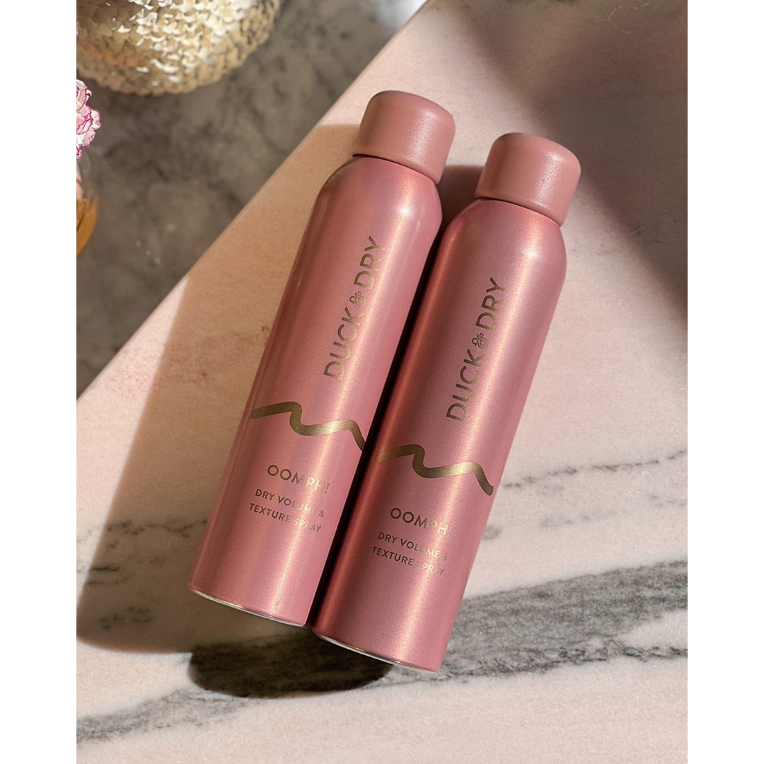 Two pink metallic cans of Duck & Dry Oomph! Dry Volume & Texture Spray with gold lettering, lying on a pink and grey marble surface in sunlight.