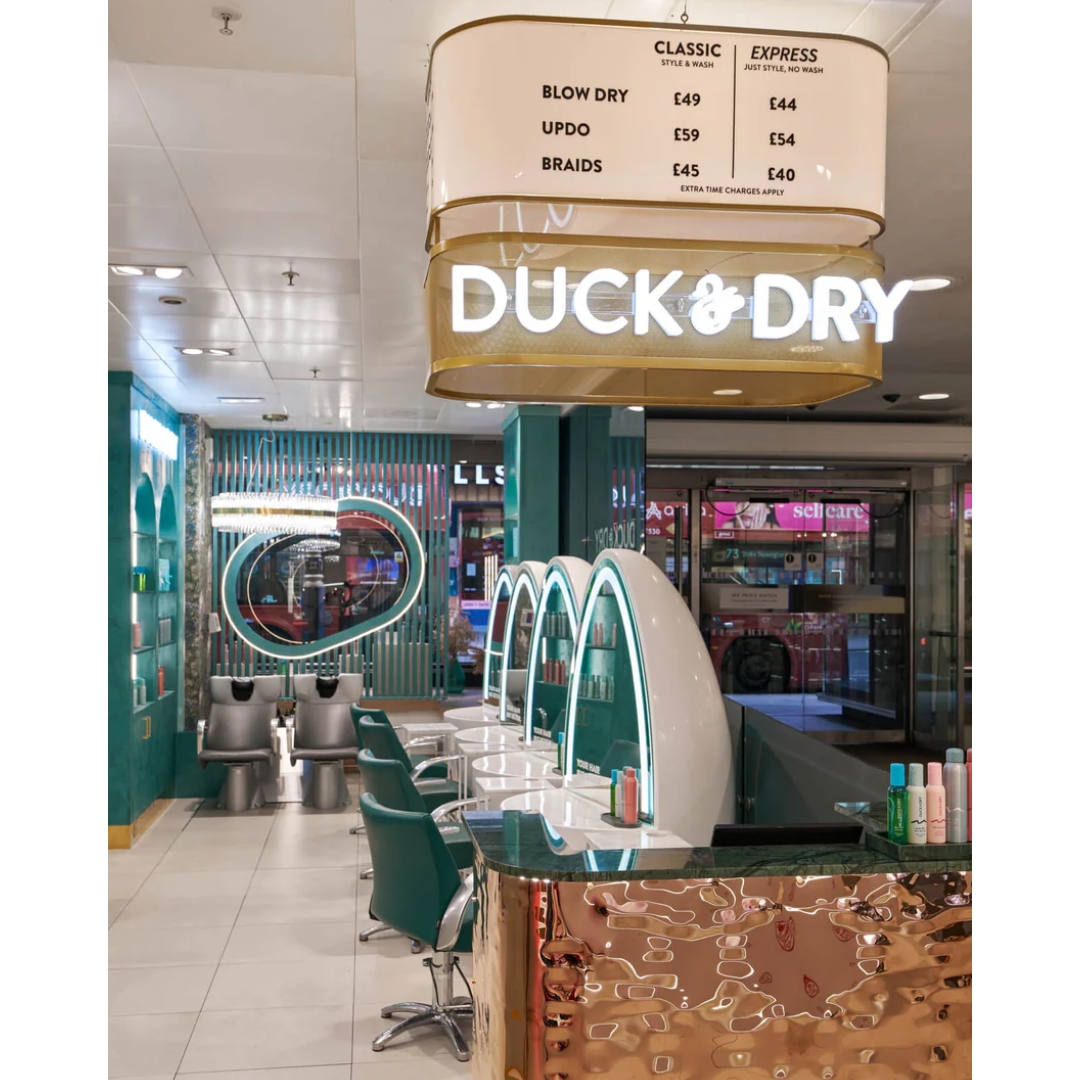 Interior of Duck & Dry’s Oxford Circus salon, with emerald-green styling chairs, white arched mirror stations, a rippled gold reception desk, and an illuminated sign listing blow-dry, updo, and braid prices.