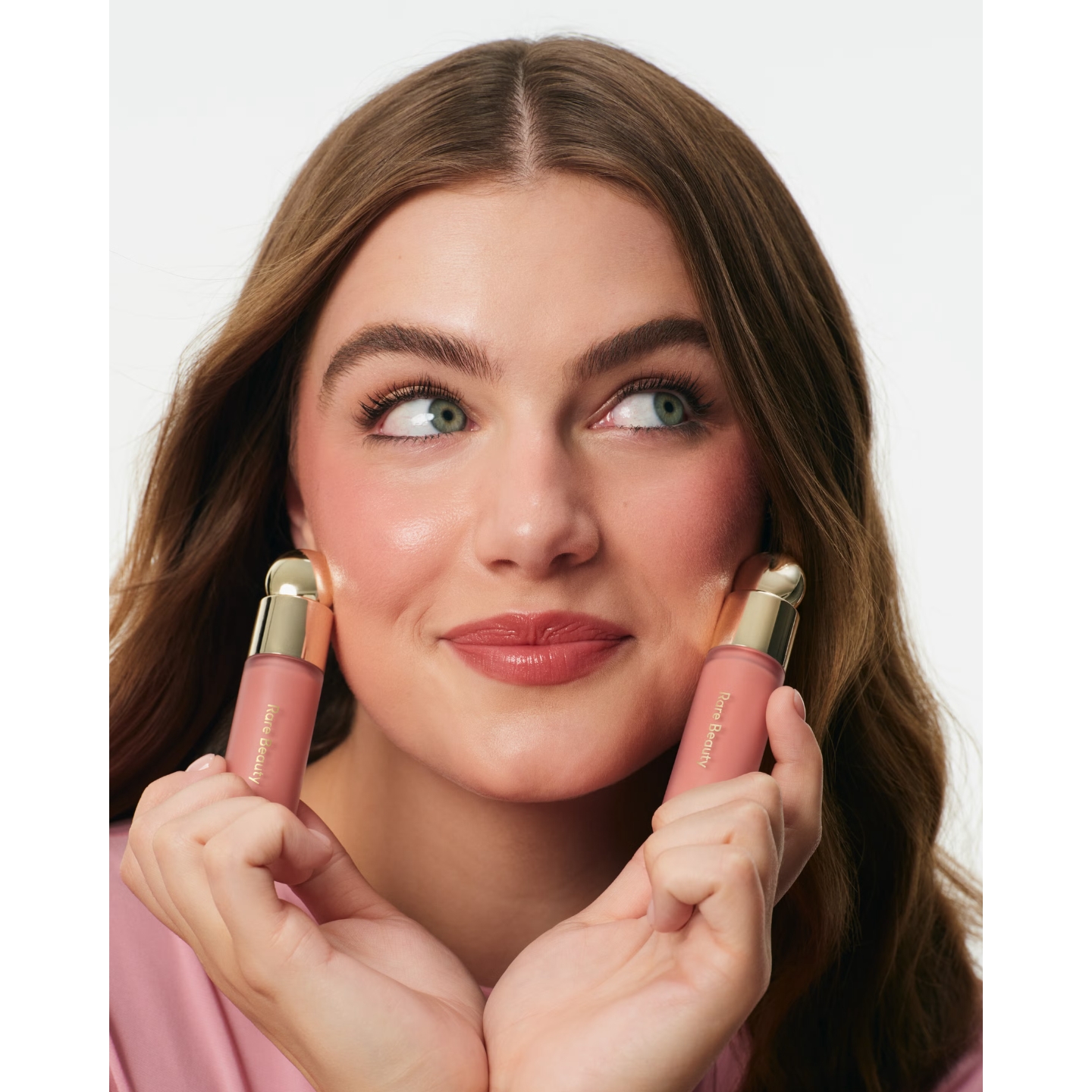Actress Ella Bright smiling playfully while holding two tubes of Rare Beauty Soft Pinch Liquid Blush to her cheeks.