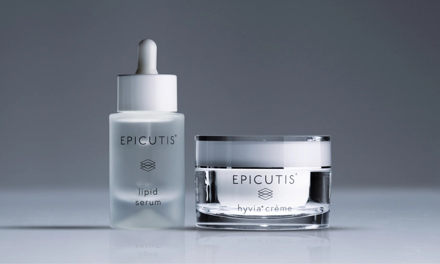 A frosted glass dropper bottle of Epicutis Lipid Serum sitting beside a clear jar of Epicutis Hyvia Crème against a cool gray background.