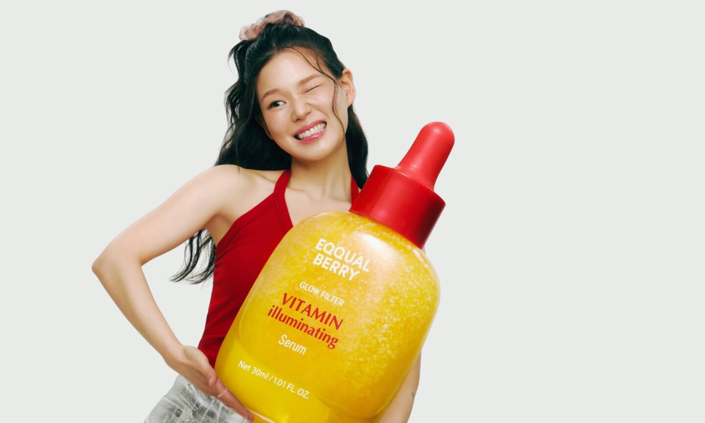 A model winking while holding a giant replica of the yellow Eqqualberry Glow Filter Vitamin Illuminating Serum bottle, featured in the brand's UK launch announcement on Beauty News Daily.