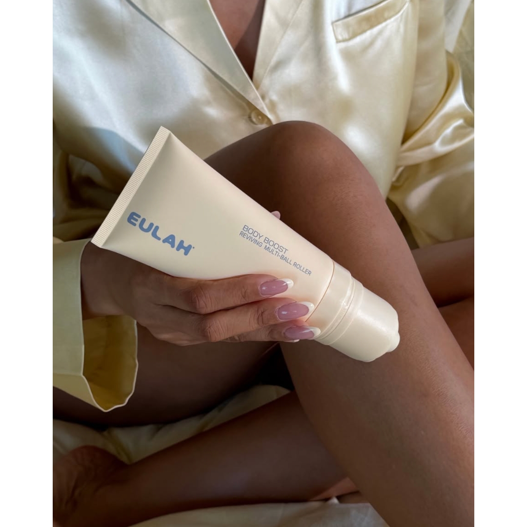 A person wearing a pale yellow silk robe rolling the Eulah Body Boost Reviving Multi-Ball Roller tube along their leg.