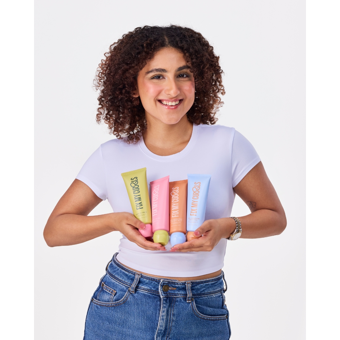 Fix My Curls founder Anshita Mehrotra, smiling and wearing a white cropped T-shirt and jeans, holds the brand’s haircare products against a white background
