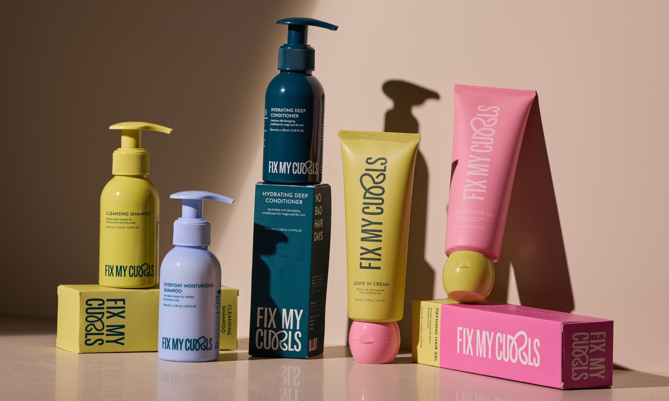 Fix My Curls Cleansing Shampoo, Everyday Moisturising Shampoo, Hydrating Deep Conditioner, Leave In Cream, and Defining Hair Gel arranged on their boxes against a warm beige backdrop, with soft shadows