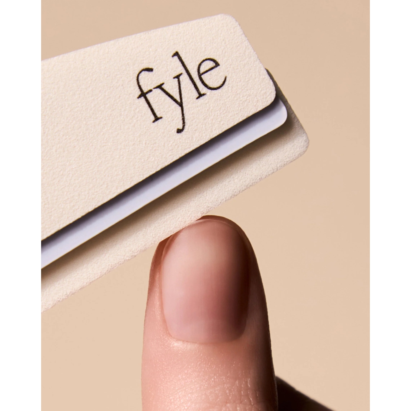 Extreme macro close-up of a natural fingernail resting directly beneath a cream-colored Fyle nail buffer showing the brand logo on a neutral background.