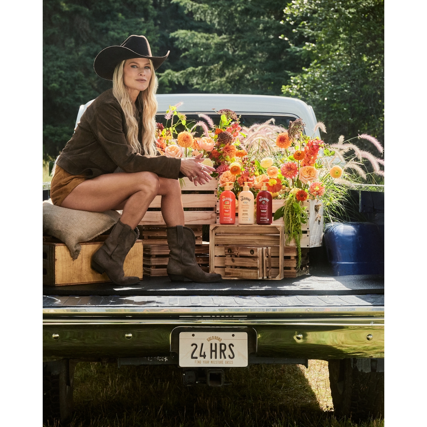 Actress Ali Larter wearing a cowboy hat, brown jacket, and western boots sitting on the bed of a pickup truck alongside crates of vibrant flowers and Gold Bond Scented Body Lotions in Island Peach, Vanilla Dream, and Cherry Crush.