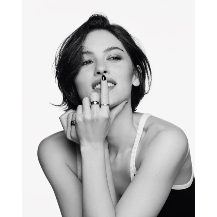 A black and white close-up of Gracie Abrams holding her index finger to her lips while wearing multiple Chanel Coco Crush rings, featured in her Chanel brand ambassador announcement on Beauty News Daily.