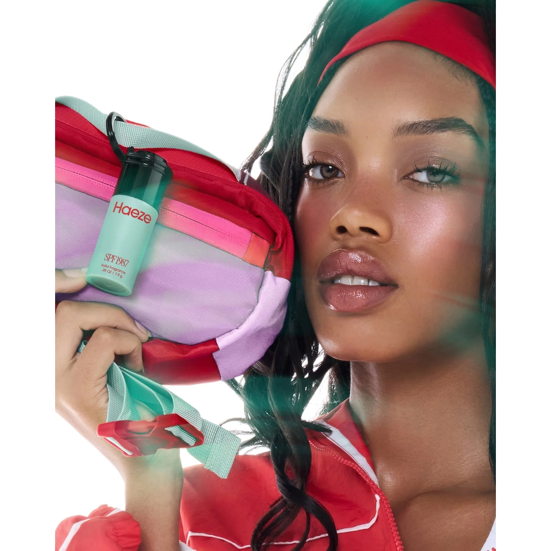 A model holds a colourful bum bag to her cheek, with a Haeze SPF 1987 Solid Fragrance clipped to its strap, in hazy, light-streaked campaign shot.