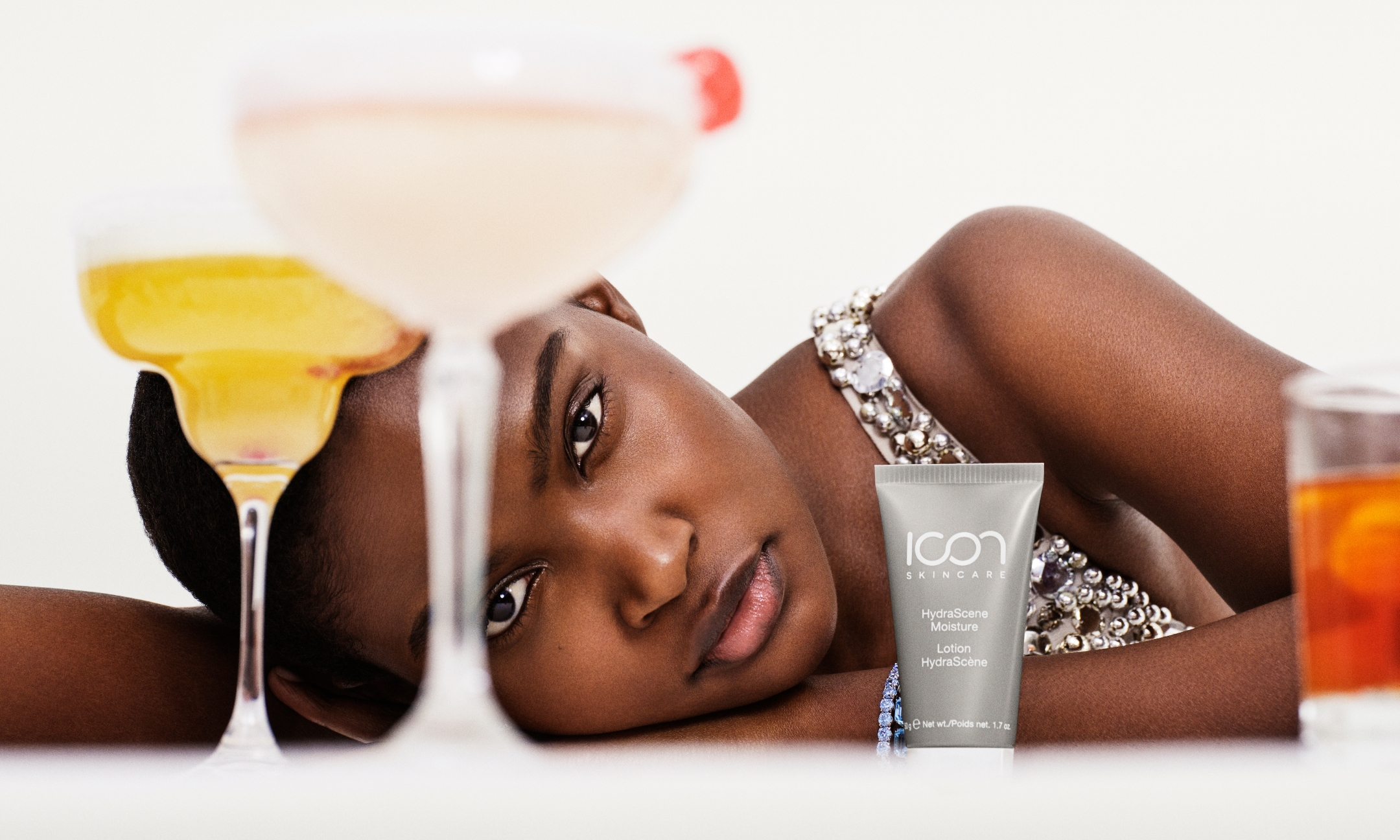 Model in an embellished top resting her head on a surface next to a tube of Icon Skincare HydraScene Moisture, framed by cocktail glasses in the foreground.