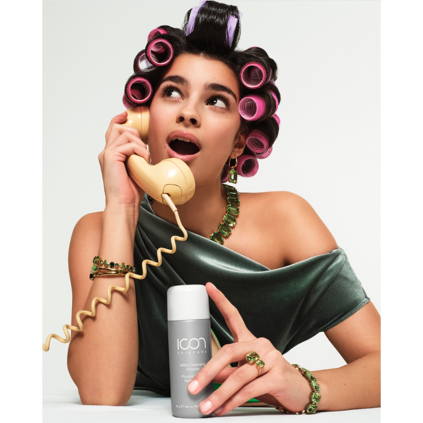 Model wearing hair rollers and green jewelry talking on a yellow rotary telephone while holding a bottle of Icon Skincare MilkIt Cleansing Powder against a plain white background.