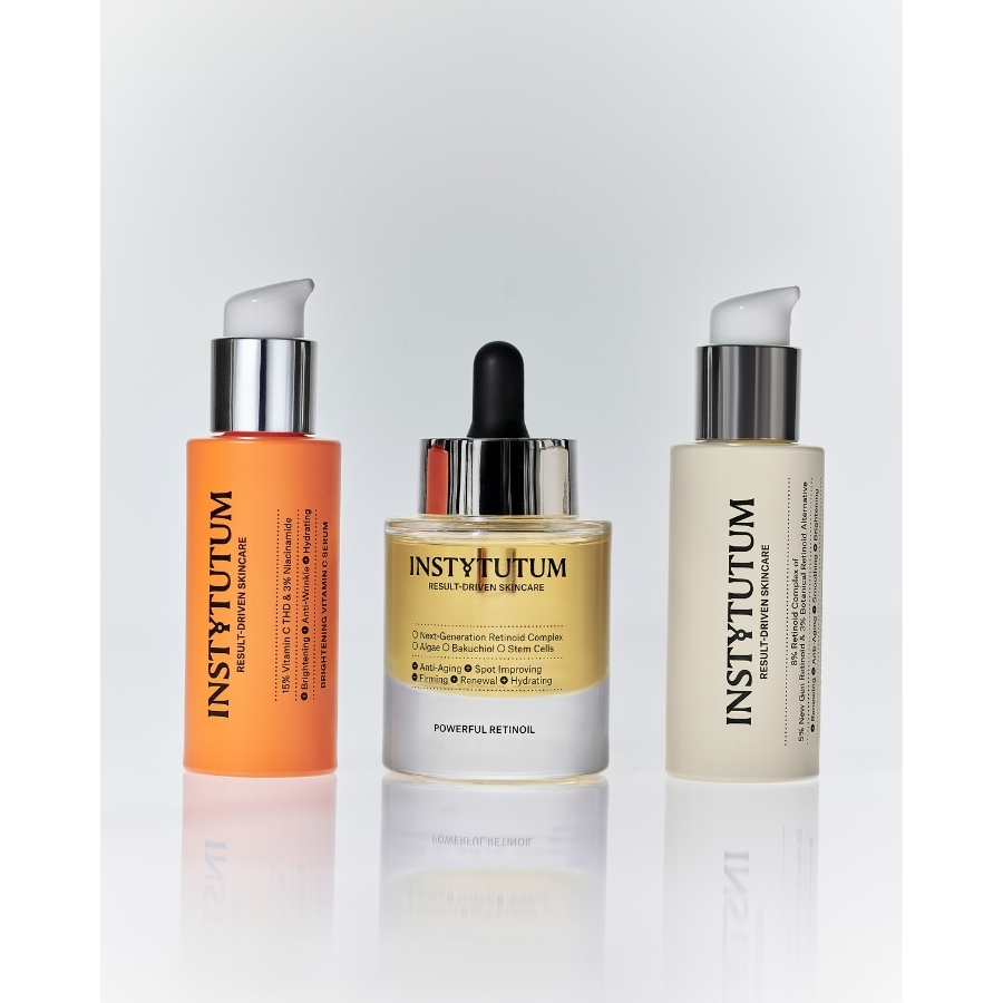 Three Instytutum serum bottles standing in a row, featuring the Brightening Vitamin C Serum, Powerful Retinoil, and Retinol Complex on a reflective white surface.