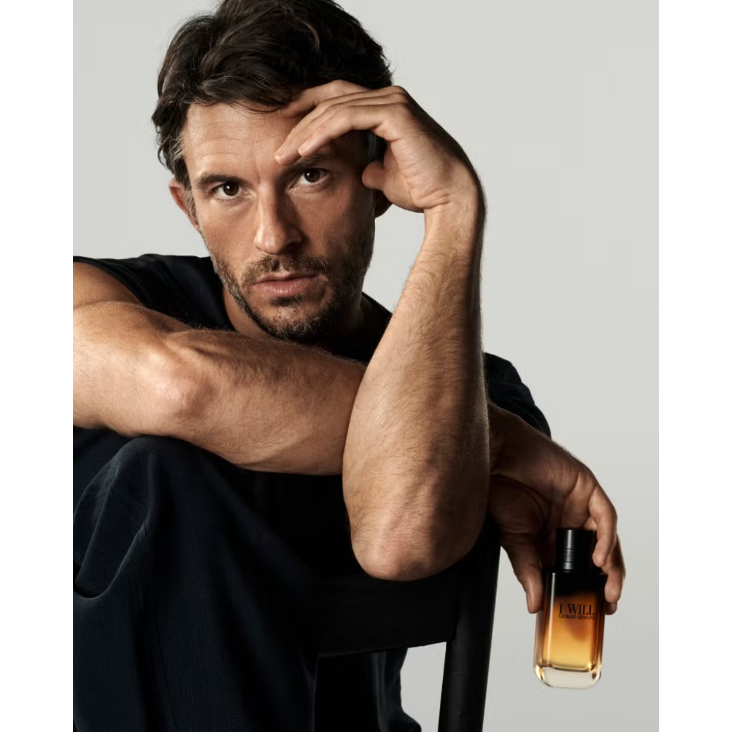 Actor Jonathan Bailey posing in a black top resting his hand against his forehead while holding a bottle of Giorgio Armani I Will fragrance against a plain backdrop.