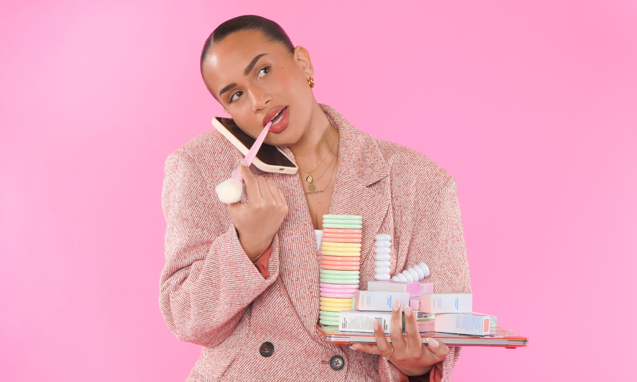 Jordana Ticia Cosmetics founder Jordana Franks holding a phone to her ear and a pink makeup brush to her lip while balancing stacked Jordana Ticia products on a laptop against a bright pink background.