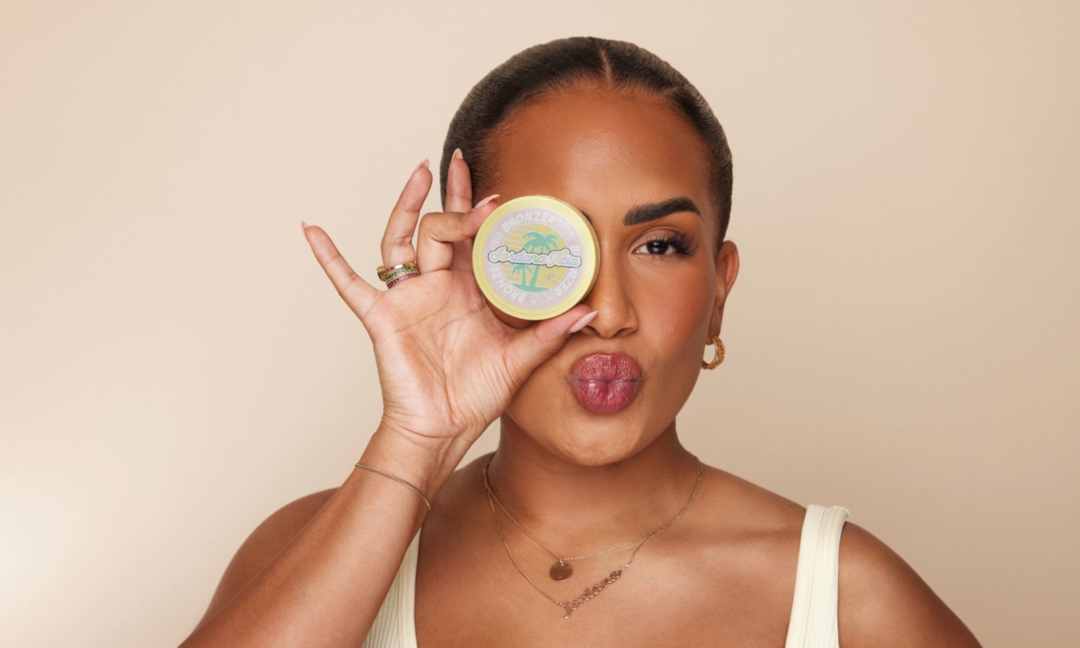 Jordana Ticia Cosmetics founder Jordana Franks holding a yellow Bronzer Duo compact over one eye while making a kissy face against a beige background.