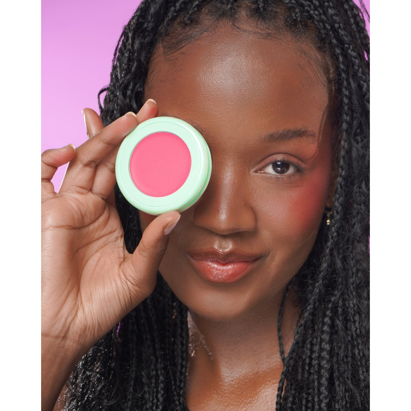 Model with braided hair holding an open mint green compact of Jordana Ticia Cosmetics Blusher Duo in Fan Girl over one eye against a pink background.