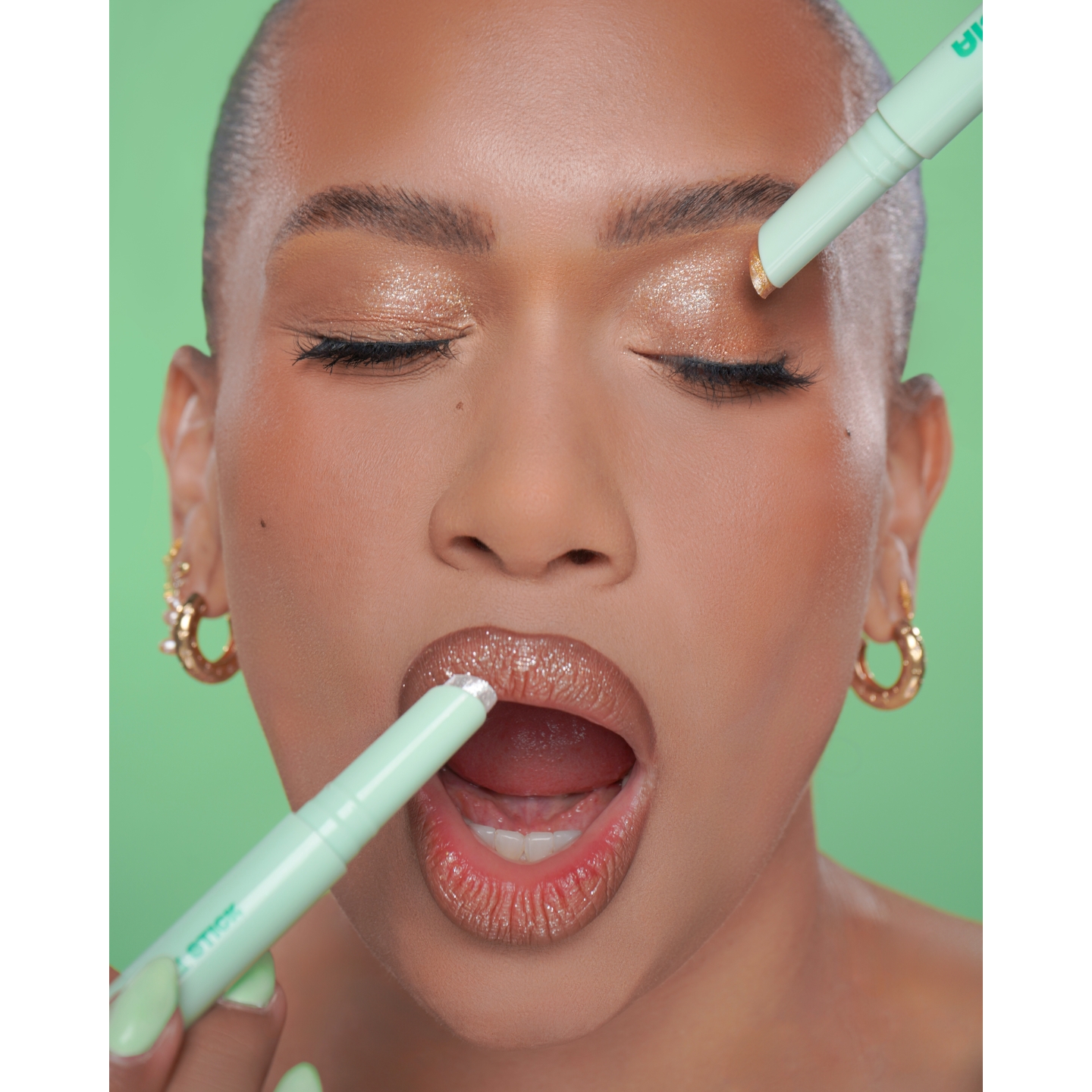 Close-up of Jordana Franks wearing shimmery eye and lip makeup applying mint green Jordana Ticia Cosmetics Gummy Twinkle Sticks to her eyelid and lips against a green background.