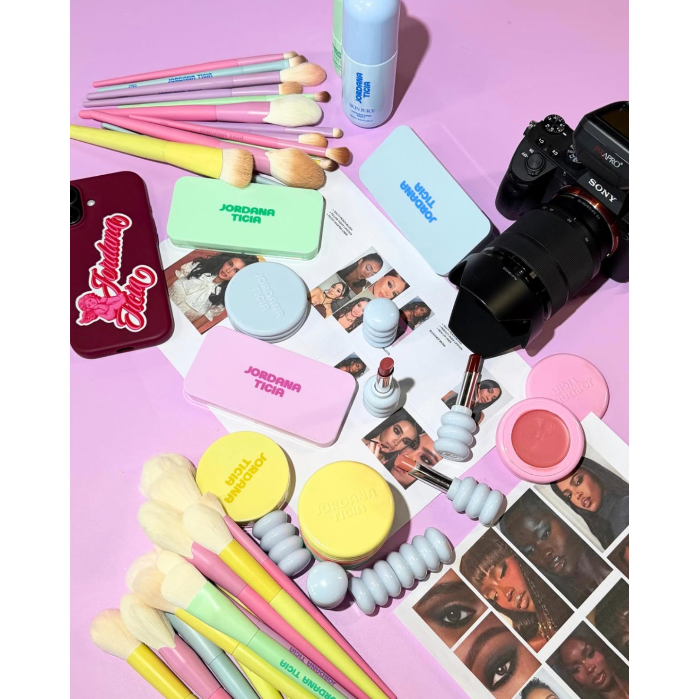 Flat lay of colorful Jordana Ticia Cosmetics products, pastel makeup brushes, Compacts, and lipsticks arranged on a purple surface next to a camera and smartphone.